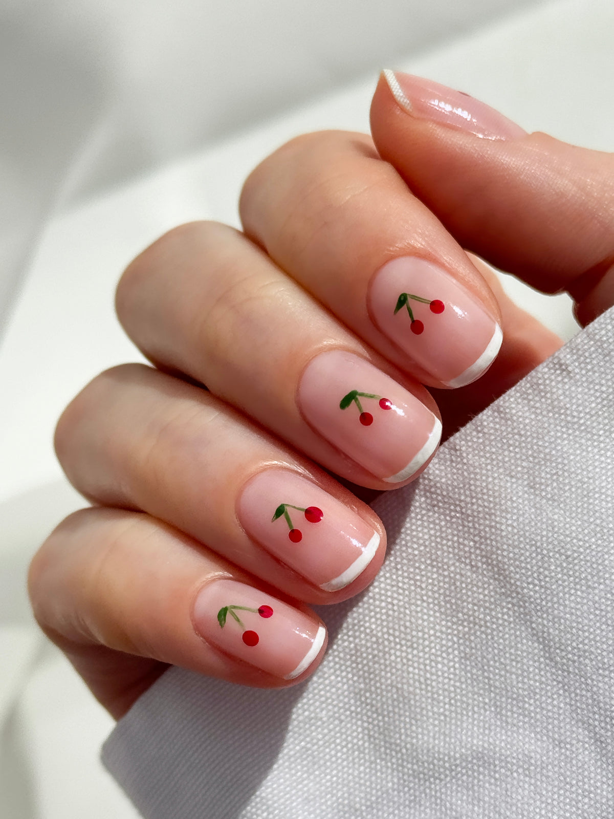 A hand with natural, glossy nails featuring small, red cherry designs and white French tips, holding a piece of white fabric.