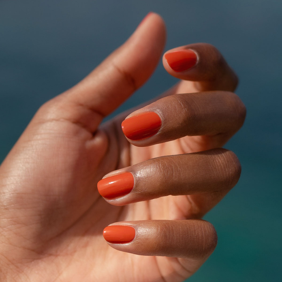 Gros plan sur une main à la peau mate aux ongles parfaitement manucurés, vernis avec le Color Refresher orange brillant de Manucurist pour prolonger la tenue de la manucure, sur un fond bleu flouté.
