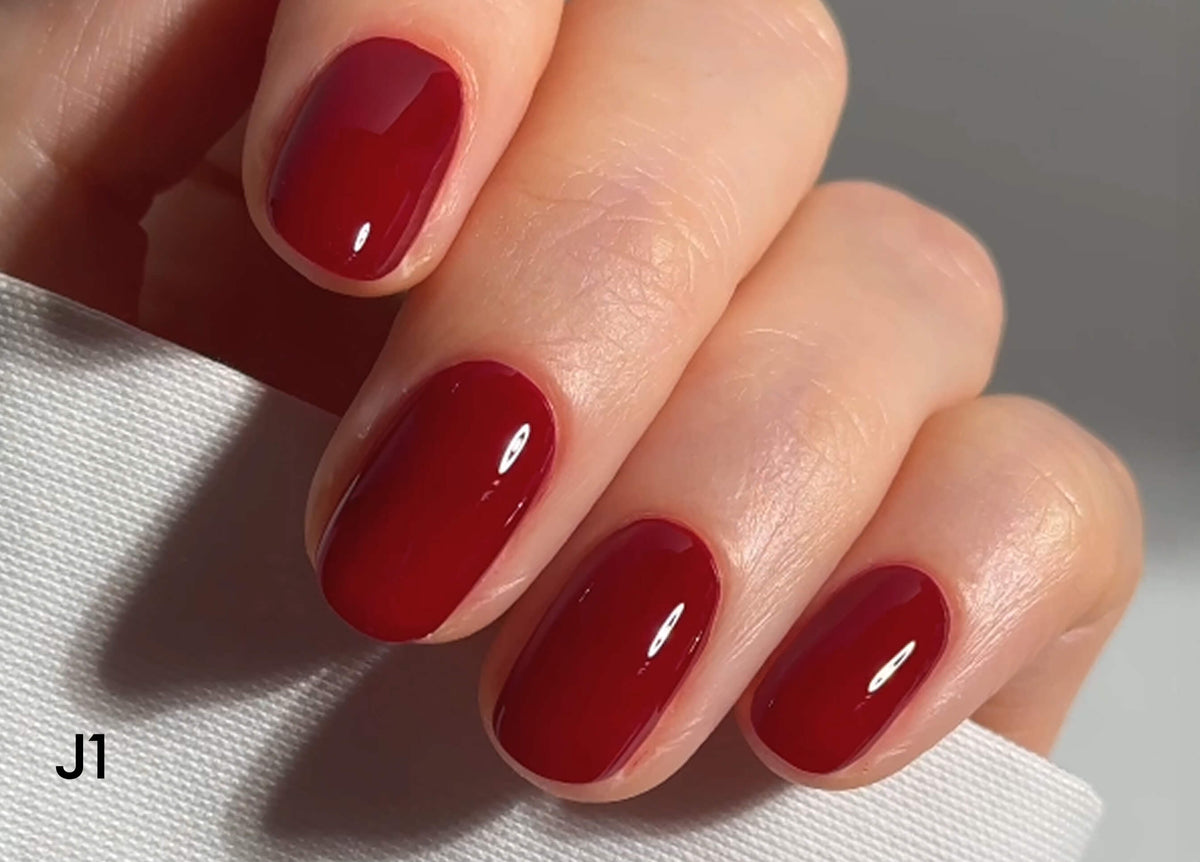 A close-up of a hand with neatly manicured nails painted in glossy deep red polish, holding a white textured fabric. The nails are oval-shaped and well-groomed. The bottom left corner has the text J1.