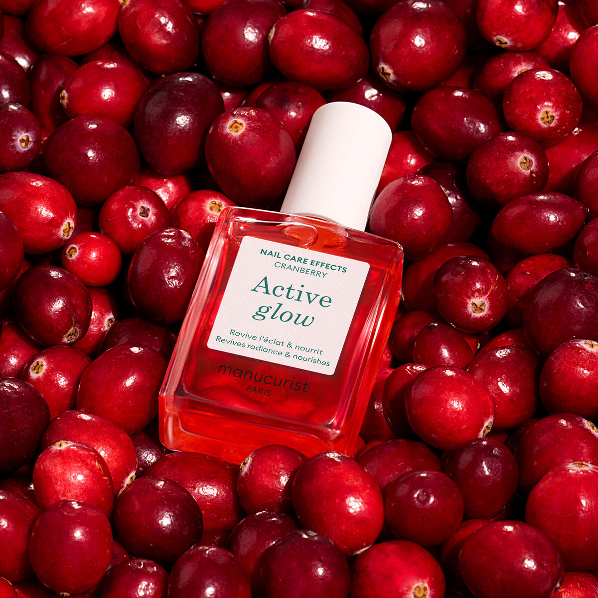 A square bottle of Manucurist Active™ Glow Cranberry nail care oil, enriched with plant-based ingredients, is displayed on a bed of fresh cranberries. The label highlights cranberry and boasts benefits such as reviving and nourishing nails, making it ideal for achieving a healthy, tinted glow.