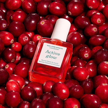 A square bottle of Manucurist Active™ Glow Cranberry nail care oil, enriched with plant-based ingredients, is displayed on a bed of fresh cranberries. The label highlights cranberry and boasts benefits such as reviving and nourishing nails, making it ideal for achieving a healthy, tinted glow.