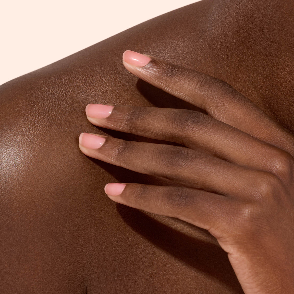 A close-up of a hand with neatly manicured nails painted in Manucurist Active™ Glow Cranberry, the vibrant polish shining against smooth, dark brown skin as it rests gently on a bare shoulder, all set against a light beige background.