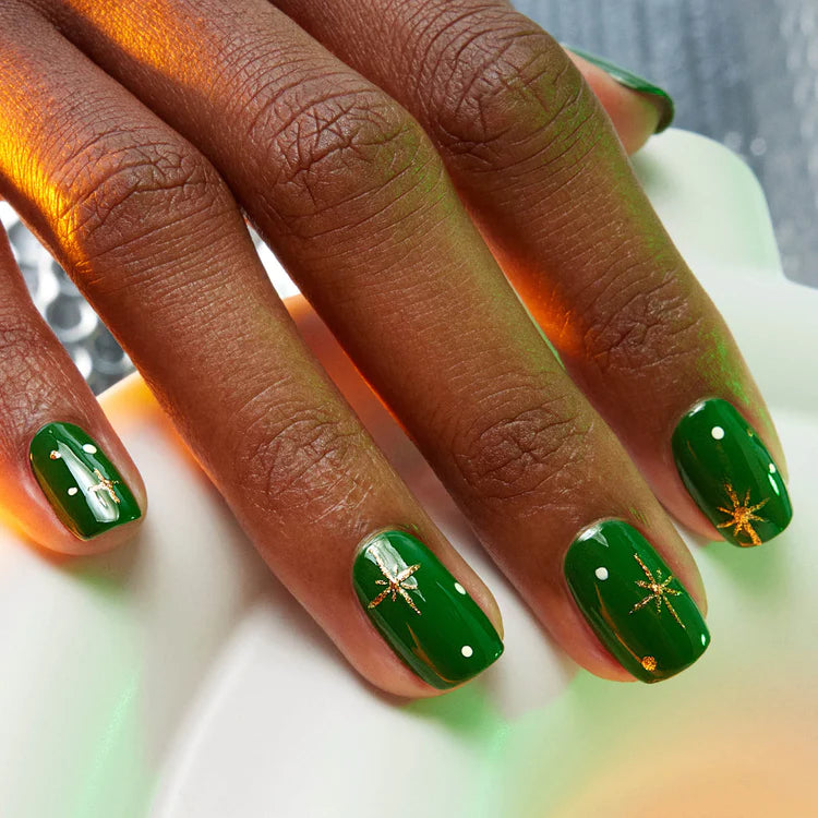 A hand with green nail polish, featuring small white dots and gold starburst designs on some nails, resting on a white surface.