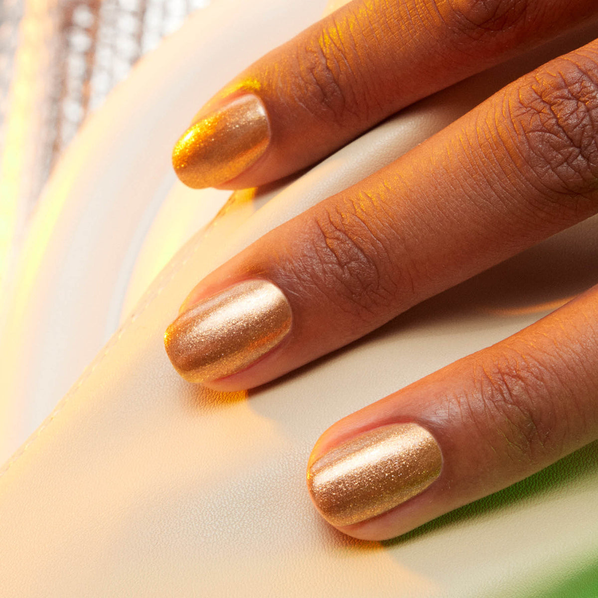 A close-up of a hand resting on a white surface, with fingernails painted in Manucurist’s Shimmer, a shiny honey-glazed bronze nail polish.