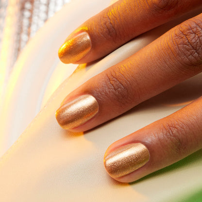 A close-up of a hand resting on a white surface, with fingernails painted in Manucurist’s Shimmer, a shiny honey-glazed bronze nail polish.