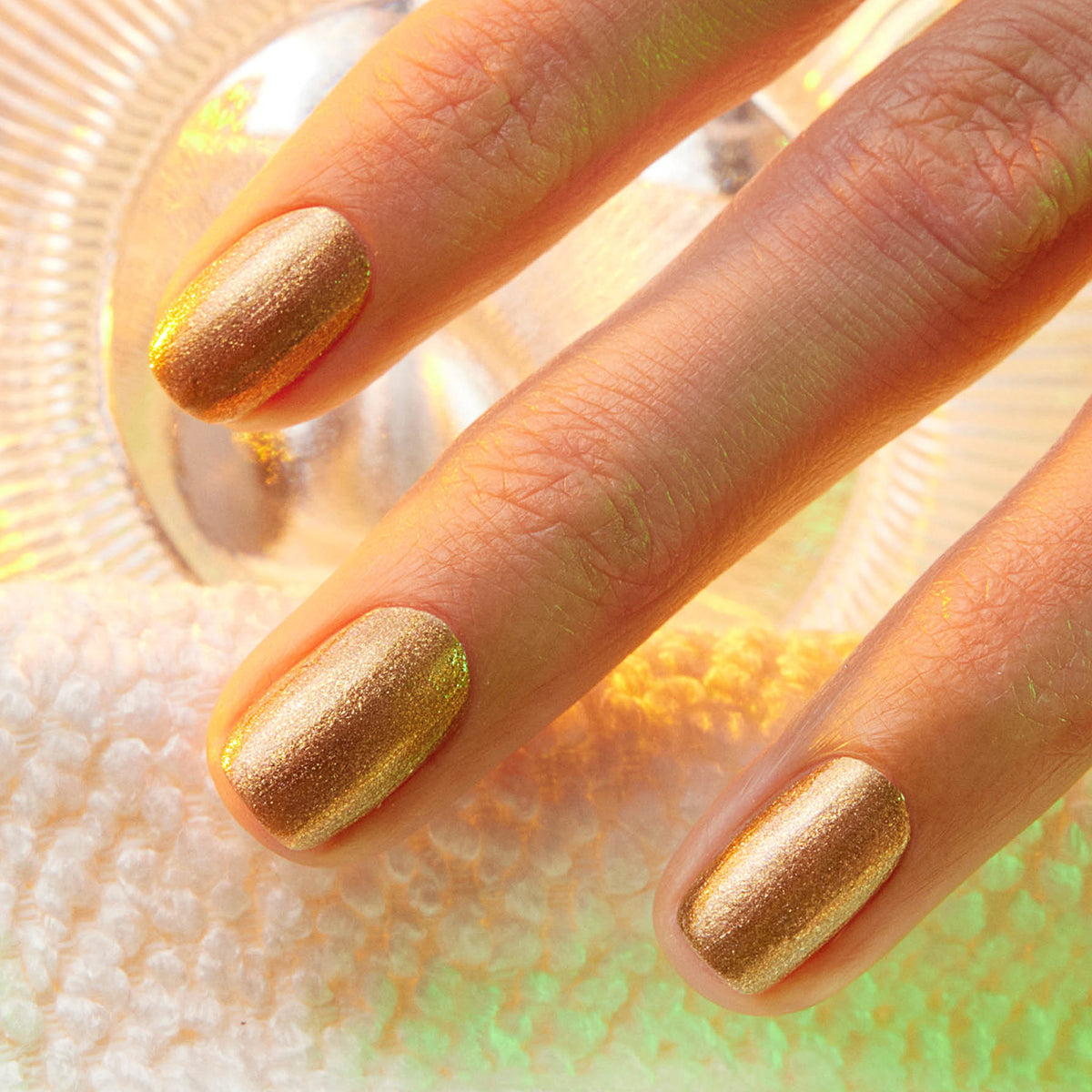 Three neatly manicured fingers showcase Manucurist’s Shimmer nail polish—a cool, shimmery gold—resting on a textured white surface with a glass object in the background.