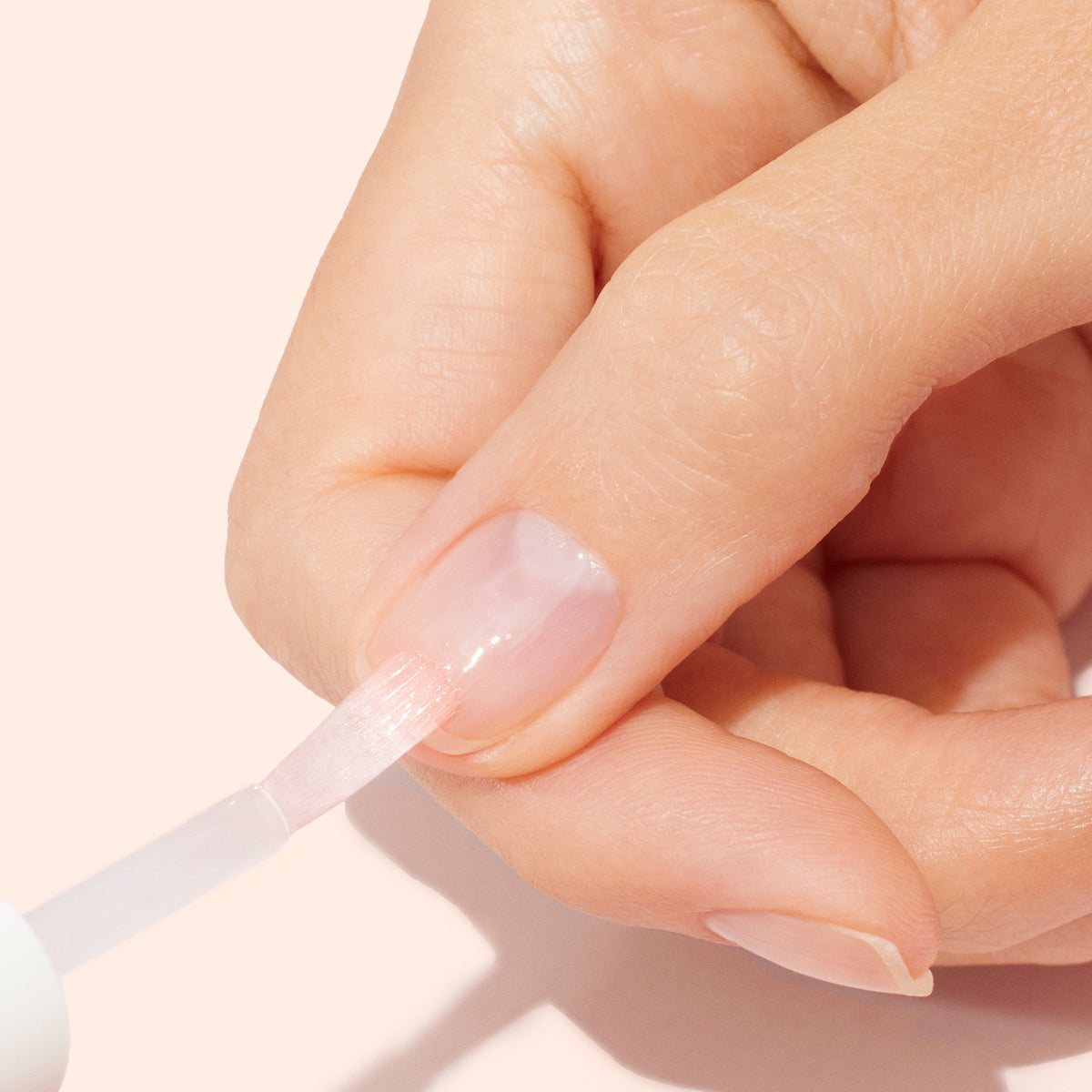 A close-up of a hand applying Manucurist S.O.S. Nail Mask to fingernails with a brush against a light pink background.
