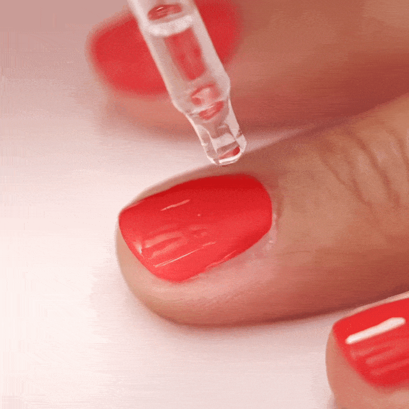 A close-up of a person’s hand with bright coral-red nail polish being applied with a clear liquid dropper onto a fingernail, likely as a finishing coat or nail treatment.