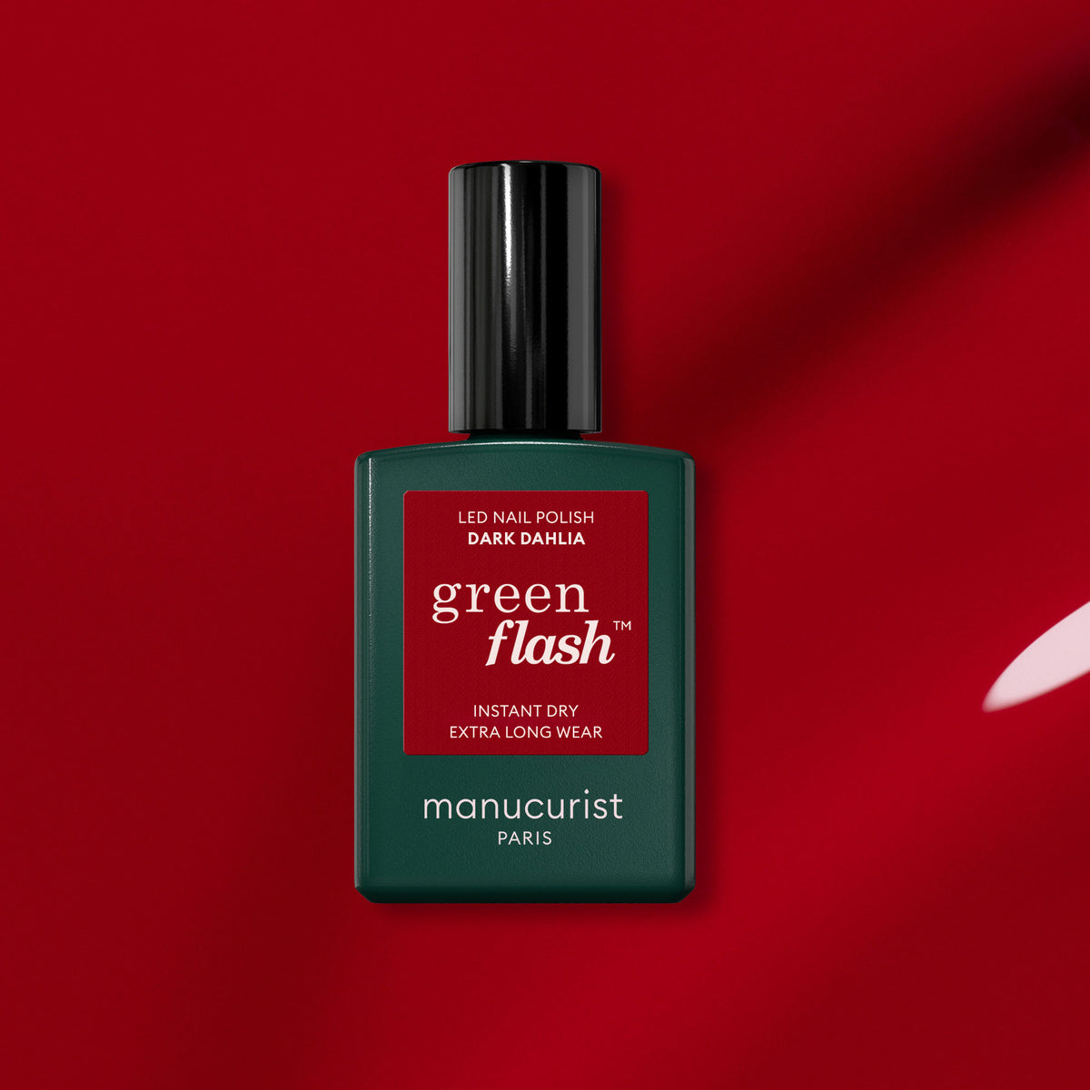 A bottle of Manucurist Dark Dahlia gel polish sits on a red background. The green bottle with a black cap and red label showcases the rich hue of the Dark Dahlia shade.