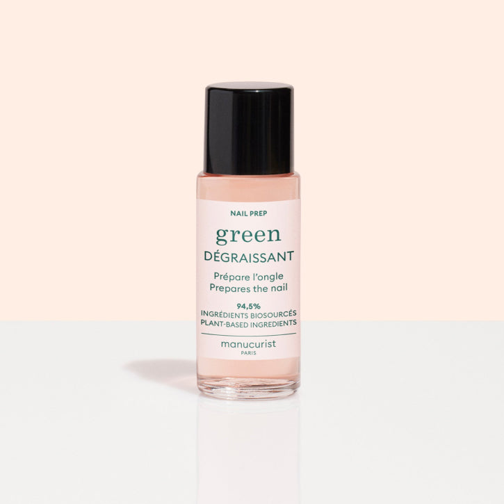 A bottle of Manucurist Paris Green Nail Prep Degreaser with black cap, clear liquid inside, and green and pink label, set against a two-tone pale pink and white background.