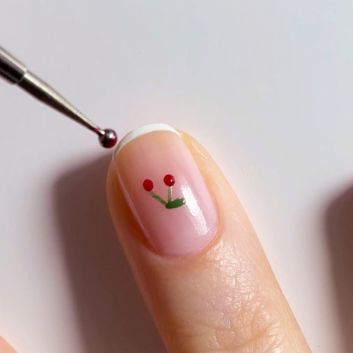 A close-up of a fingernail with a minimalist cherry nail art design, featuring two red dots connected by green stems, and a tool applying a red dot to the nail.
