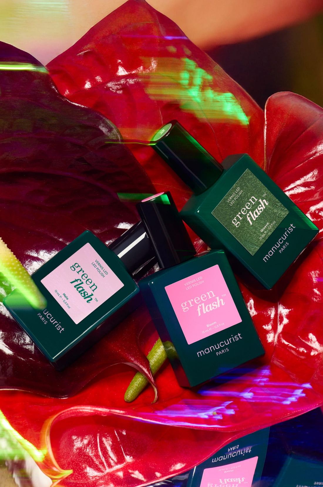 Three bottles of Green Flash nail polish by Manucurist Paris are arranged on glossy red anthurium leaves, with vibrant, colorful lighting creating an artistic and vivid display.