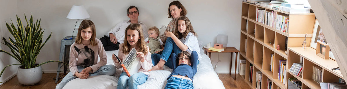 A family of seven, including adults and children, sits together on a white bed in a cozy, well-lit bedroom with bookshelves, a plant, and various books in the background.