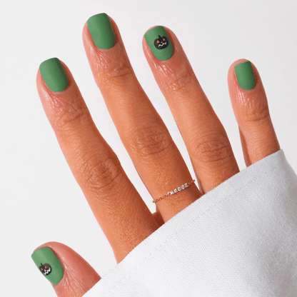 A hand with Pumpkin Patch matte green gel polish by Manucurist, featuring small black cat faces on the pinky and thumb. The look is finished with a thin gold ring and a white long-sleeve shirt—perfect subtle Halloween nail art.
