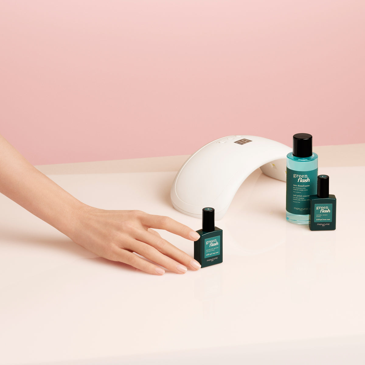 Une main attrape un flacon de vernis Green Flash Welcome kit Manucurist-test sur une surface rose clair, avec deux autres flacons et une lampe à ongles blanche à proximité sur fond rose.