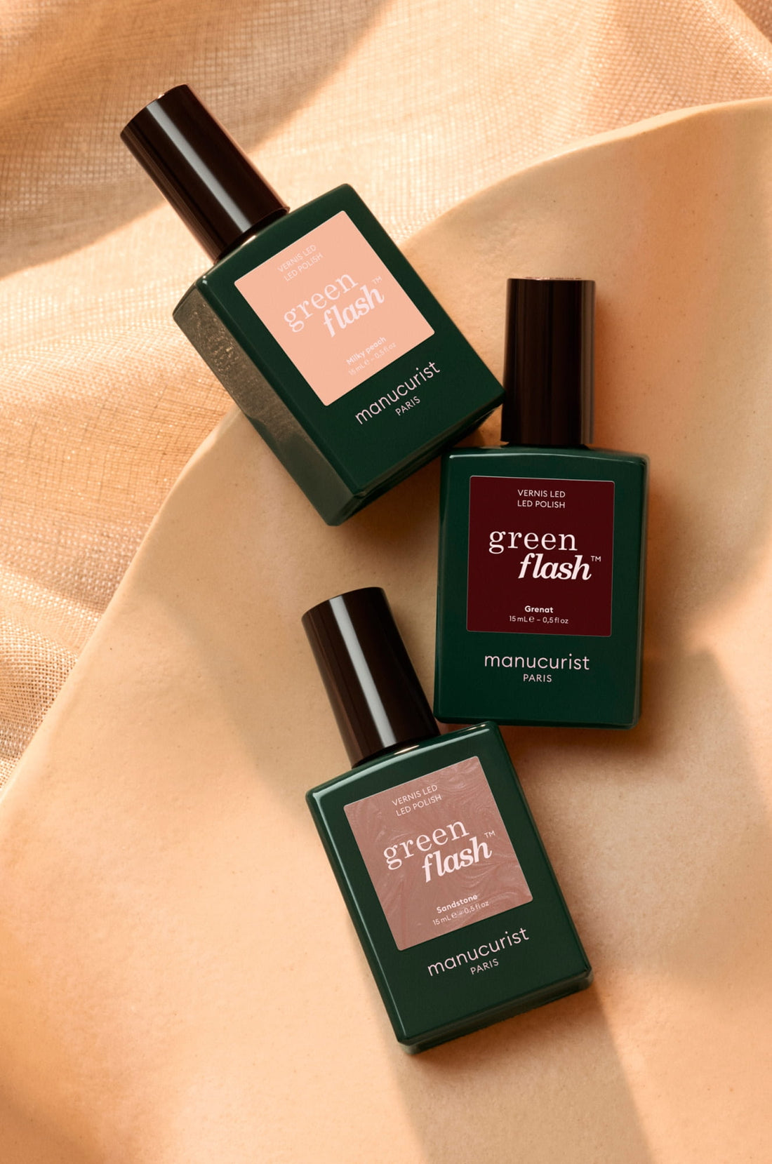 Three bottles of Manucurist Paris Green Flash nail polish are arranged on a beige surface, with green rectangular bottles and black caps. The background has a soft, textured golden fabric.