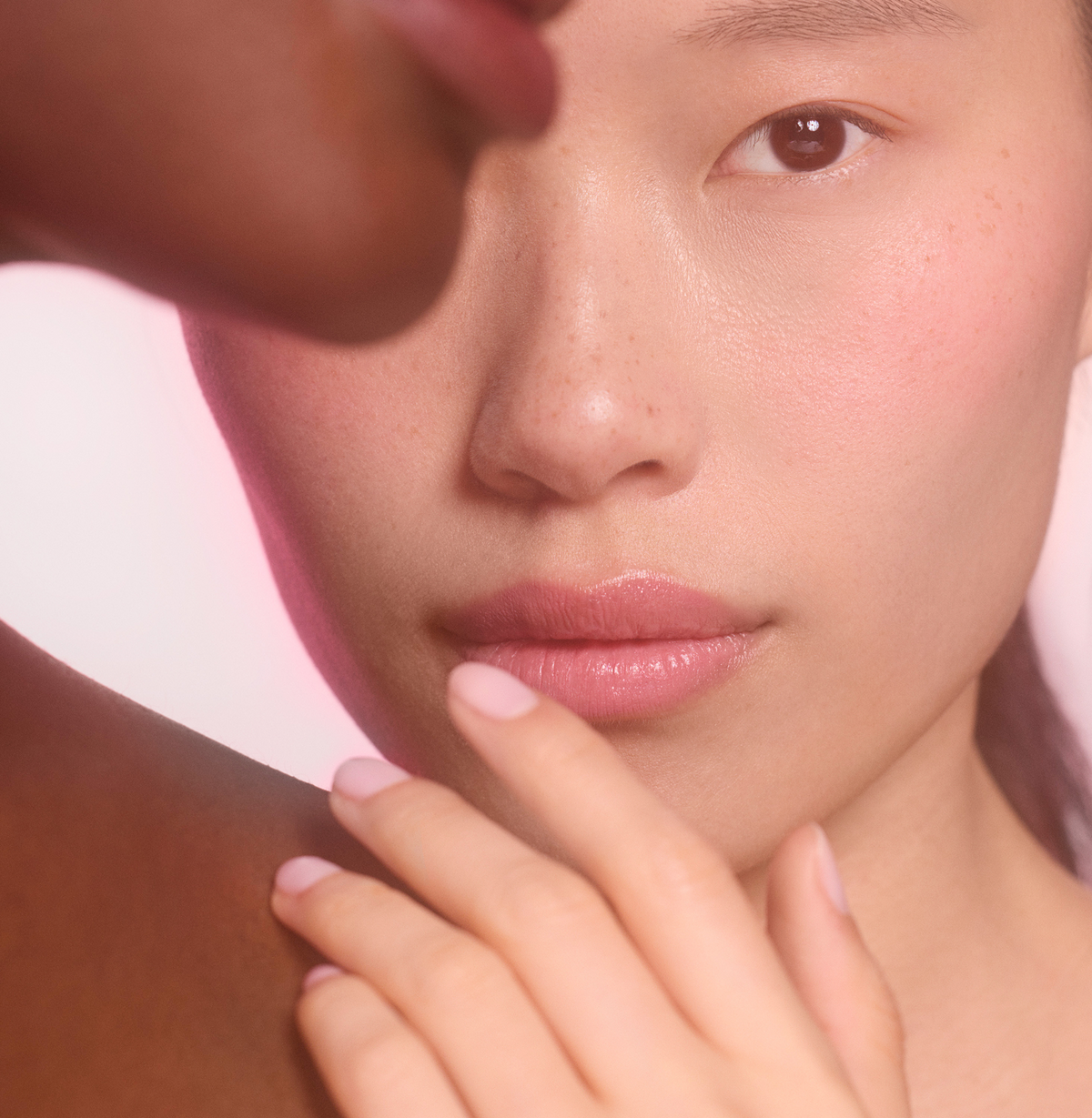 A close-up of a person with smooth, clear skin looking at the camera, while another persons face and shoulder appear softly in the foreground.