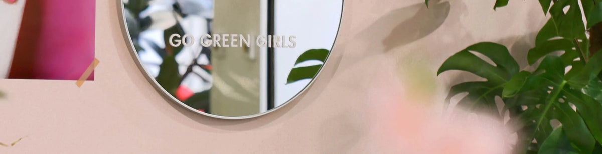 A round mirror with the words GO GREEN GIRLS etched on it is mounted on a pale pink wall, next to leafy green plants and a partially visible poster.