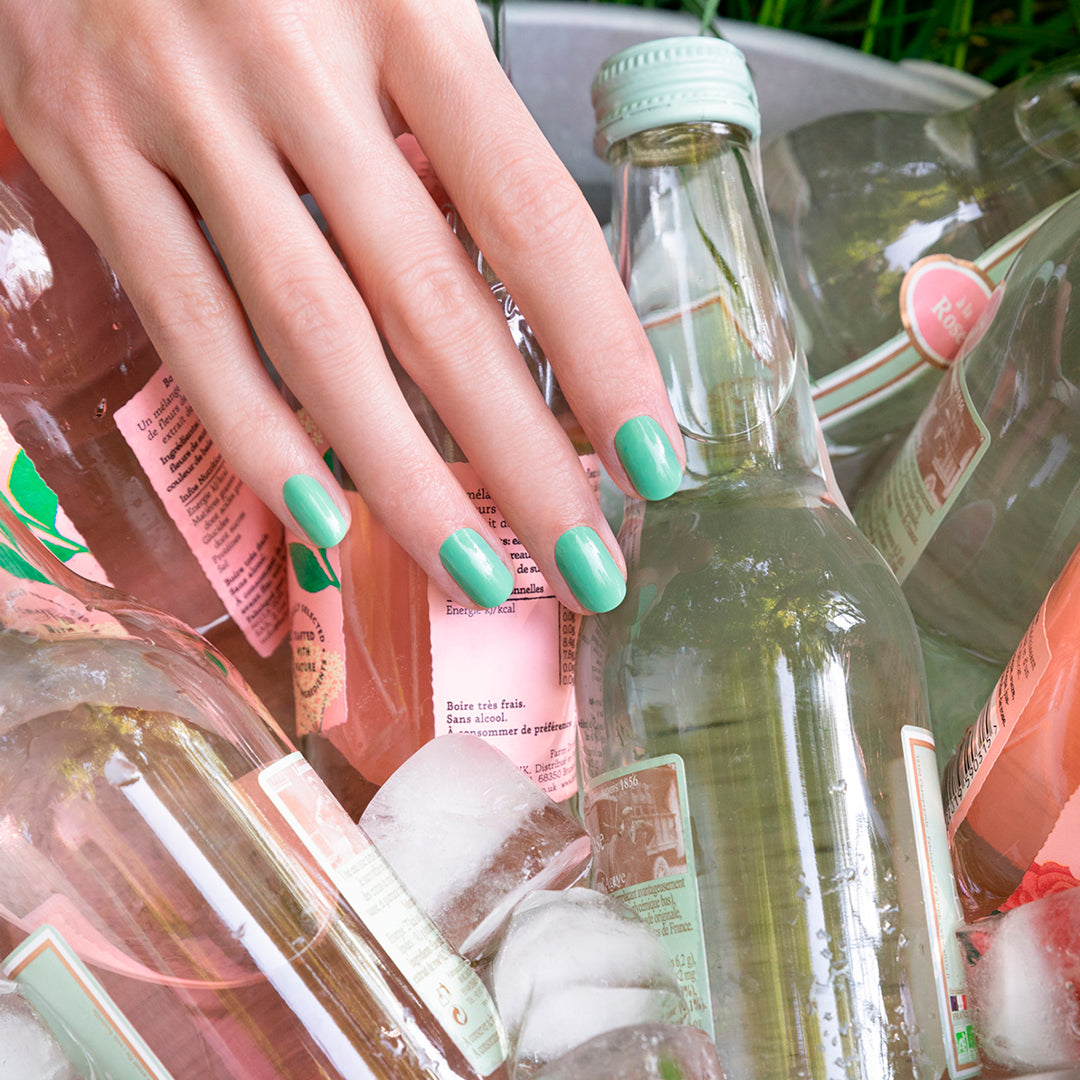 Une main avec le Set You Me Under the Sea Green Flash™ de Manucurist repose sur des bouteilles de boissons rafraîchies dans des glaçons, à l’intérieur d’un seau en métal.