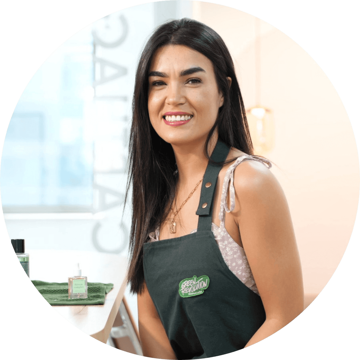 A smiling woman with long dark hair wears a black apron over a light-patterned top, sitting indoors beside a table with beauty products. She appears to be in a professional or salon setting.