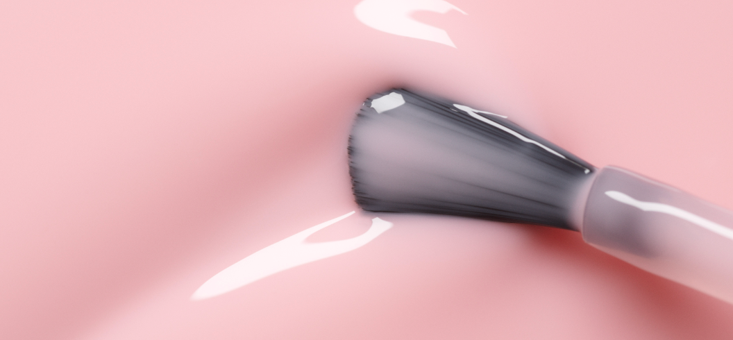 A close-up of a nail polish brush applying glossy, light pink nail polish on a smooth pink surface. The brush bristles and shiny liquid texture are clearly visible.