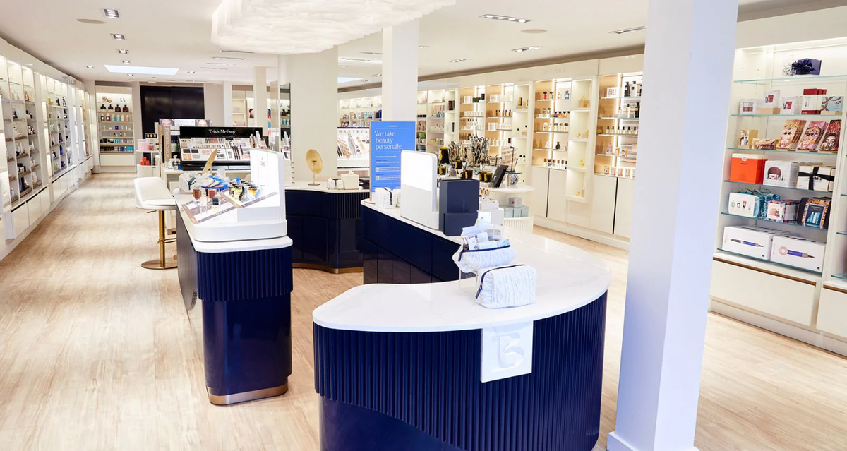 A bright, modern beauty store with white walls and light wood floors, featuring shelves filled with various skincare and cosmetic products, and sleek navy and white display counters in the center.