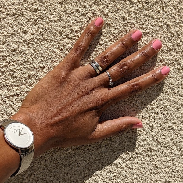 Une main aux ongles roses repose contre un mur beige texturé. La main porte deux bagues et une montre blanche au design minimaliste. La lumière du soleil projette des ombres, mettant en valeur le teint naturel de la peau.