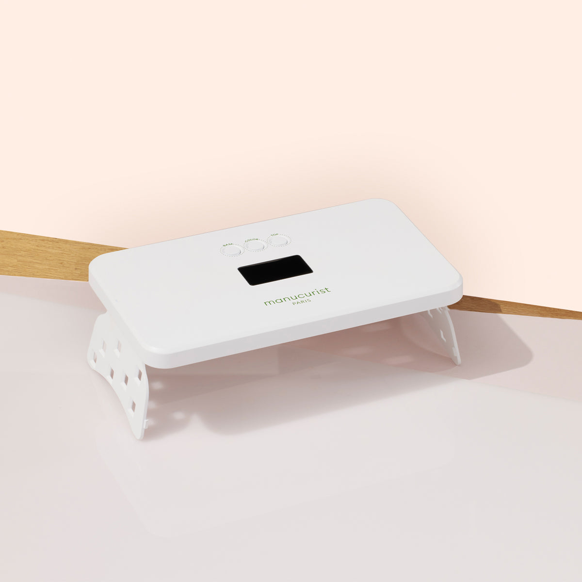 La lampe Premium 36W Manucurist Green Flash™ — une lampe LED blanche rectangulaire avec écran numérique et boutons de commande — est posée sur une surface rose clair et en bois, idéale pour sécher rapidement le vernis gel Green Flash™.