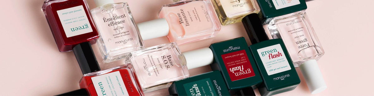 A flat lay of several bottles of nail polish in various shades and colors, arranged diagonally on a light pink background. The bottles have square shapes and bold, colorful labels.
