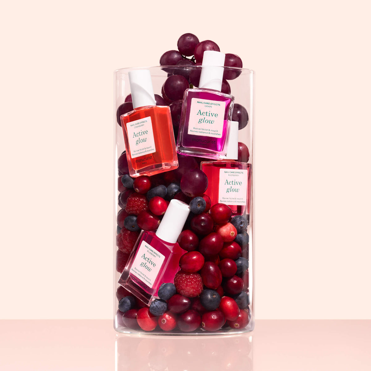 A Manucurist Customizable Active™ 5-pack is displayed in a clear glass container with red grapes, cranberries, blueberries, and raspberries, creating a unique arrangement against a light pink background.