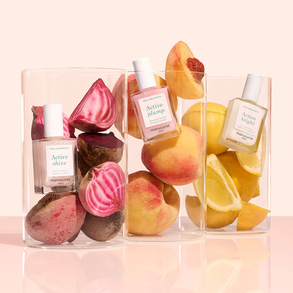 Three clear glass containers each hold a Manucurist Customizable Active 5-pack nail polish and fresh produce—beets with Active Shine, peaches with Active Plump, and lemons with Active Bright—set against a pale background.