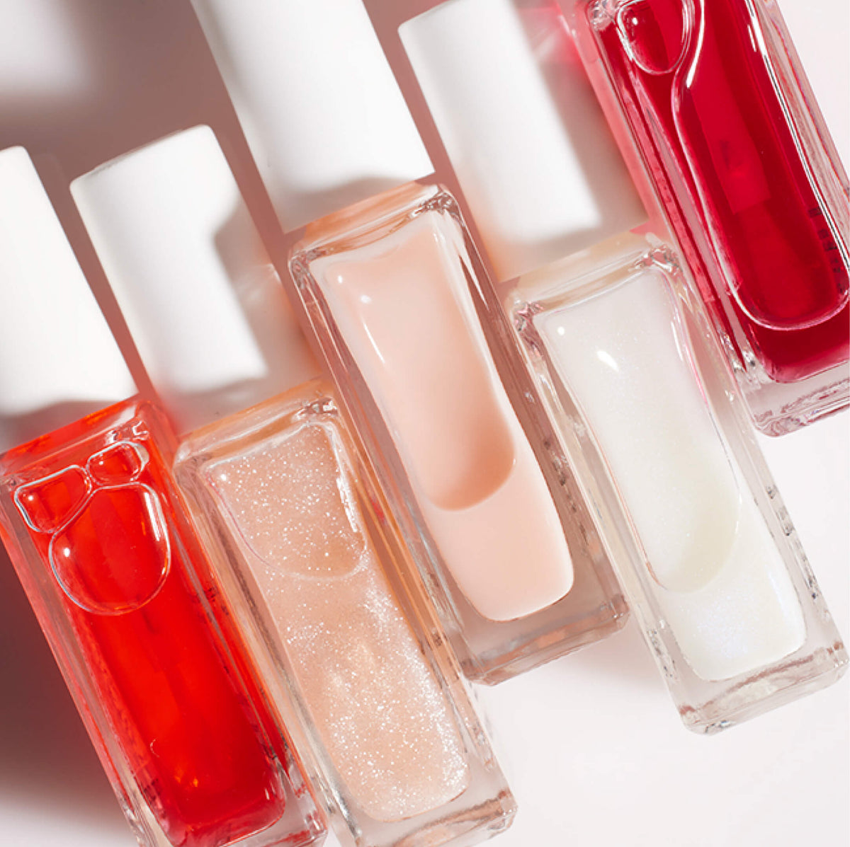 Six bottles of nail polish in various shades of red, pink, peach, and white with glossy and glitter finishes, arranged diagonally on a white surface with soft shadows.