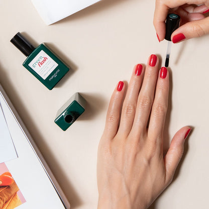 Une personne applique un Top Coat transparent du kit Welcome Manucurist-test Green Flash sur ses ongles vernis en rouge. Deux flacons de vernis à ongles et un magazine ouvert reposent sur une surface beige à côté de sa main.