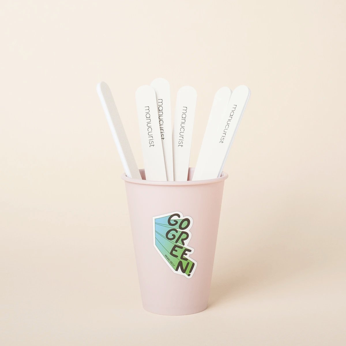 Un pot rose clair avec un sticker « GO GREEN ! » contient cinq limes à ongles blanches posées à la verticale sur un fond beige uni. Les limes à ongles portent l'inscription Manucurist.
