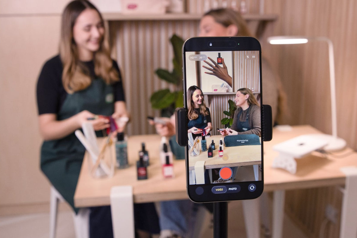 Two women sit at a table with skincare products and film themselves on a smartphone, which shows them talking and demonstrating products in a well-lit, modern room.