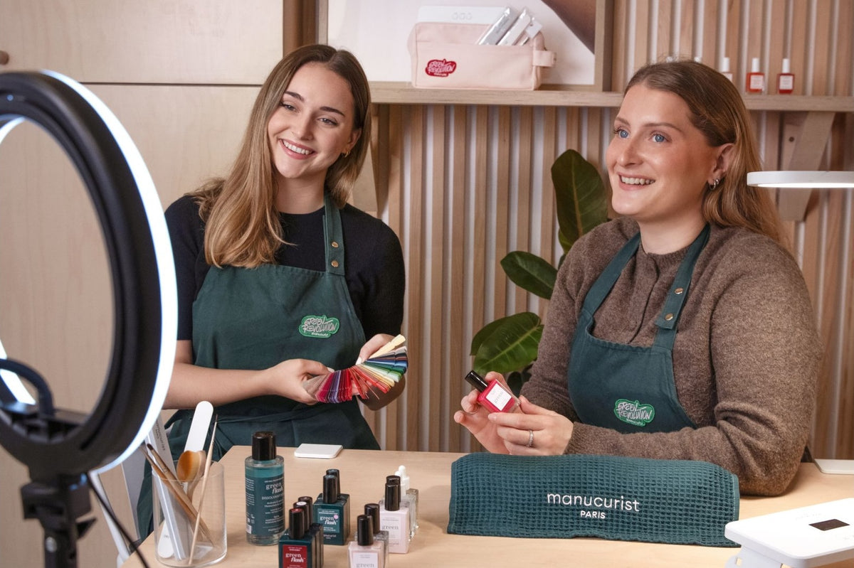 Two women wearing aprons sit at a nail salon table, smiling at a ring light camera. One holds nail polish color samples, the other holds a polish bottle. Nail products are on the table; shelves with more products are behind them.
