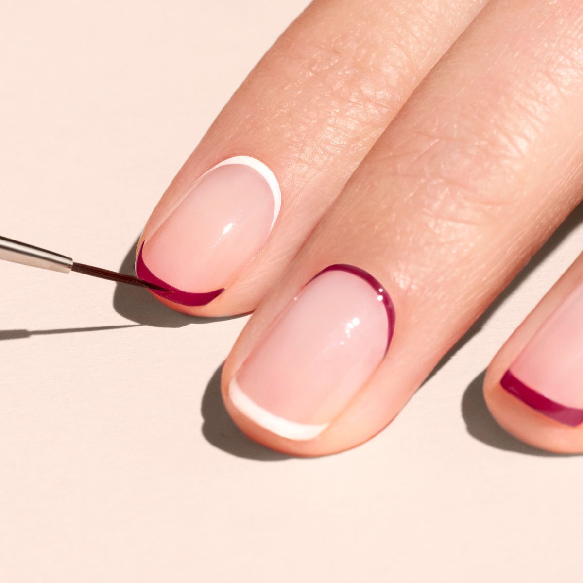 Une main aux ongles soignés arbore un nail art minimaliste ; le Liner Brush de Manucurist, aux poils synthétiques vegan, trace des courbes rose fuchsia sur une base neutre, offrant des détails de qualité professionnelle et un ongle à pointe blanche nette.