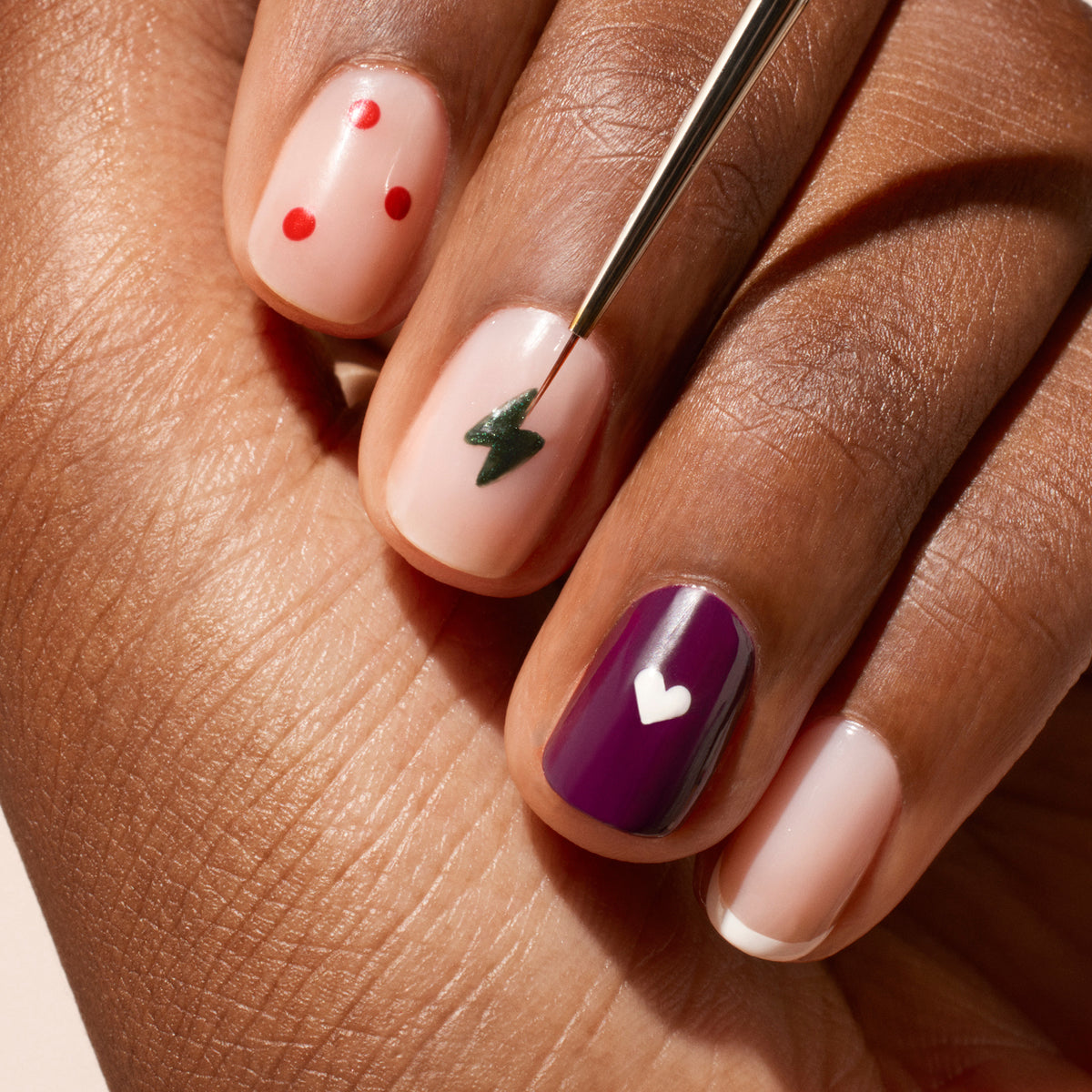 A manicured hand displays nail art—red dots, a green lightning bolt, a purple nail with a white heart, and natural nails with white tips and hearts—as the Manucurist Fine Tip Brush with vegan synthetic bristles touches one nail.