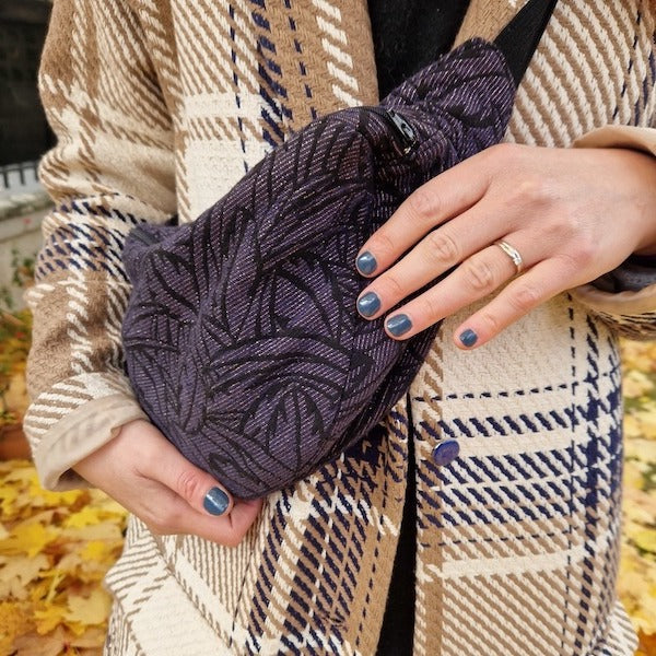 Une personne portant un manteau à carreaux avec des touches de bleu tient un sac banane violet texturé à motifs. Elle porte du vernis à ongles bleu et une simple bague en argent. Des feuilles jaunes d’automne recouvrent le sol en arrière-plan.