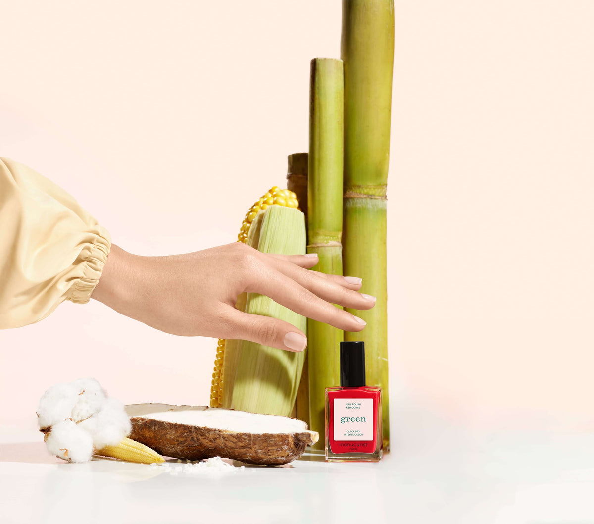 A hand reaches toward a red bottle of nail polish labeled green next to sugarcane stalks, a slice of bread with white spread, a cotton flower, corn, and a yellow beaded necklace on a pale background.