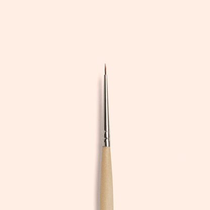The Manucurist Fine Tip Brush, made with vegan synthetic bristles, a wooden handle, and a silver ferrule, stands vertically against a light beige background—perfect for creating precise nail art.