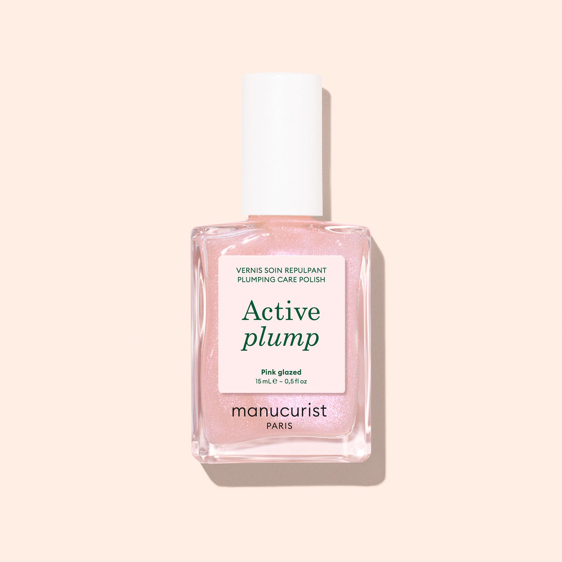 A bottle of Manucurist Active™ Plump Pink Glazed collagen nail polish in a light pink shade, featuring a white cap and displayed against a pale peach background—ideal for hydrated nails and gentle, plant-based nail care.