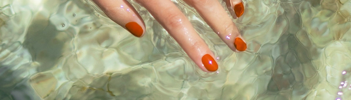 A hand with bright orange nail polish is partially submerged in clear, rippling water.