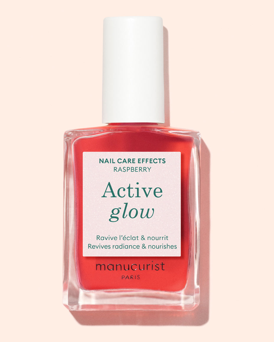 A bottle of Manucurist Paris Active Glow nail care polish in a clear rectangular glass container with a white cap, containing vibrant pink-red liquid. The label highlights its radiance-boosting and nourishing effects.
