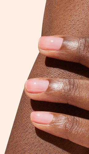 A close-up of a hand with neatly manicured, natural pink nails resting gently on a persons leg against a light beige background.