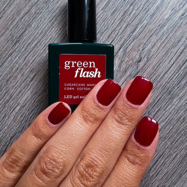 Une main aux ongles courts et soigneusement manucurés, peints en rouge profond, repose sur une surface en bois. Derrière la main se trouve un flacon foncé de vernis à ongles gel LED Green Flash™.