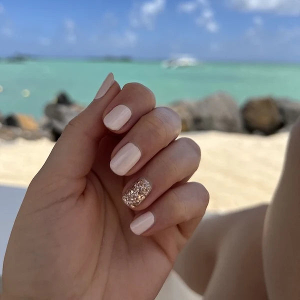 Une main aux ongles rose pâle avec un ongle pailleté en accent nail se détache sur un arrière-plan de plage tropicale avec de l'eau turquoise, des rochers, du sable et un ciel bleu nuageux.