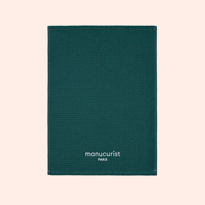 A green textured towel from Manucurist-us, featuring white "manucurist PARIS" text at the bottom center, displayed on a light beige background and ideal for surface protection.