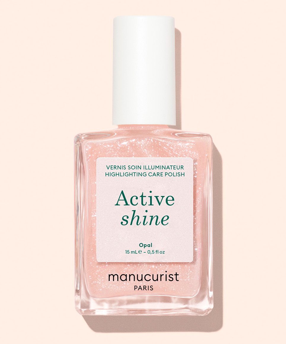 A clear glass bottle of Manucurist Paris Active Shine nail polish with a white cap, containing light pink, shimmery polish. The label highlights its nail care effects: illuminates and repairs.
