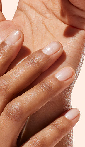 A close-up of a hand with neatly manicured, short, natural nails resting gently on an inner wrist. The nails have a glossy, light pink finish and the skin tone is medium brown.
